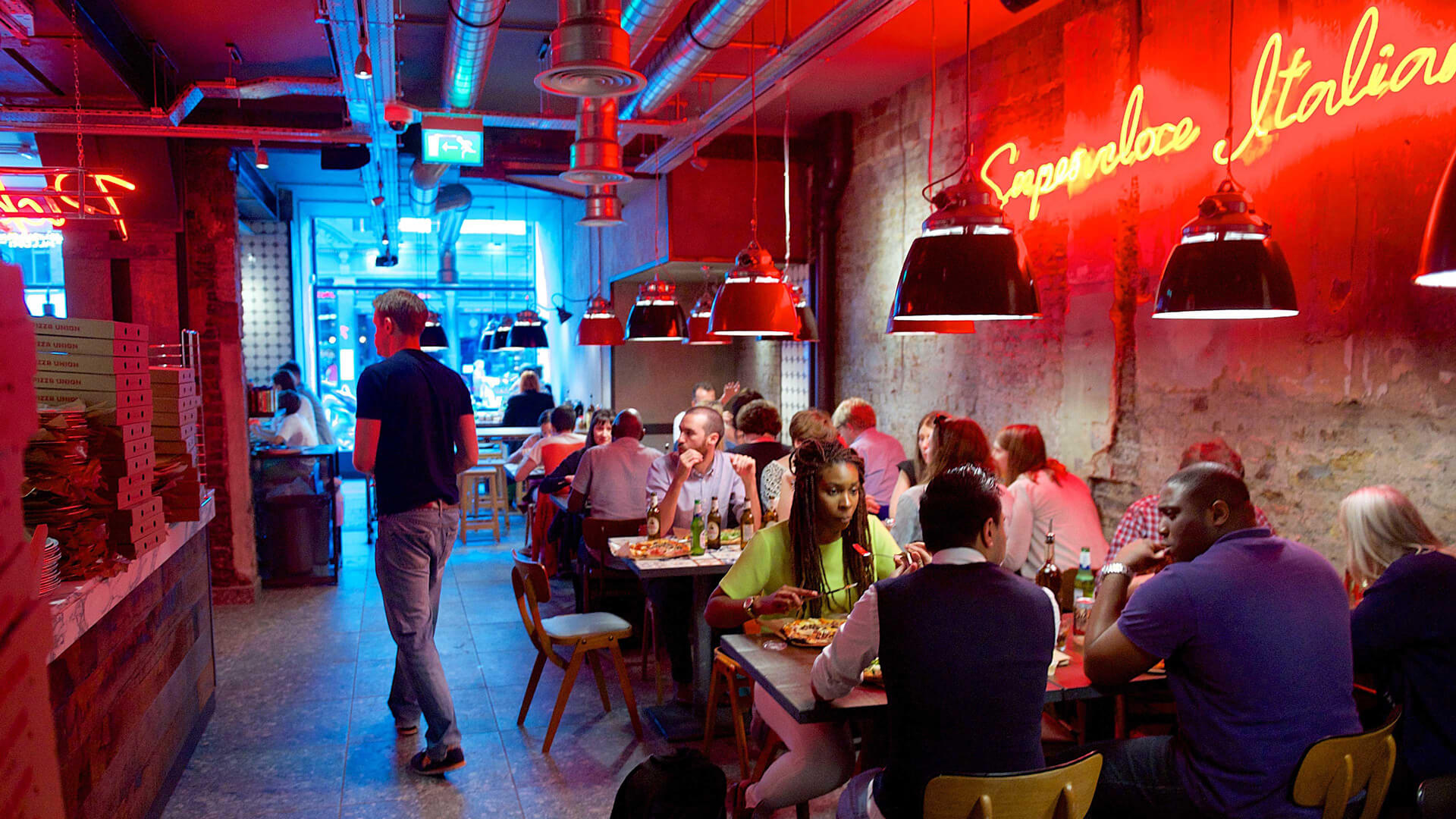Busy internal image of the Pizza union Aldgate east with rows of people sat at tables and a waitress serving pizza
